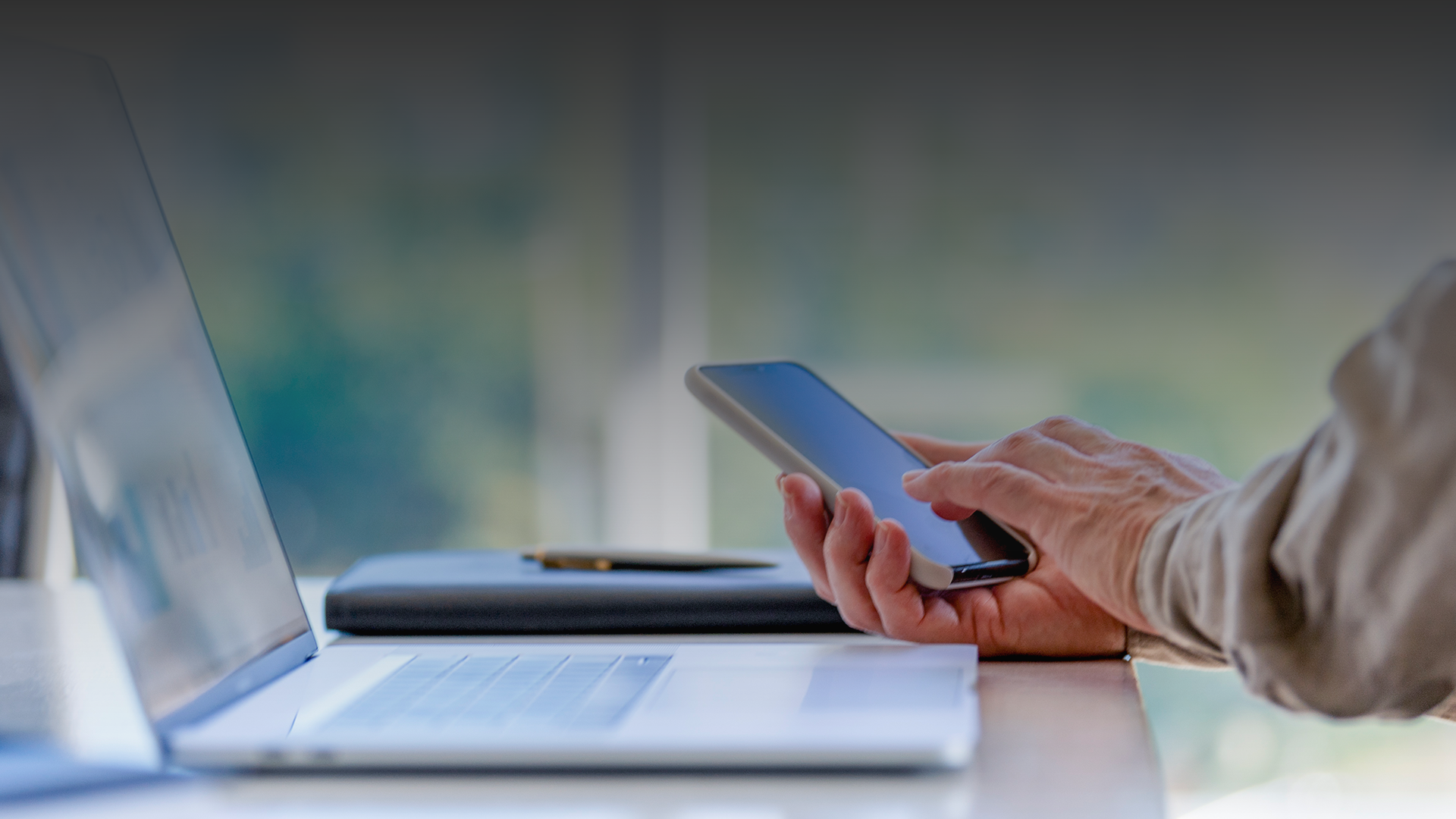 Person sitting in front of a laptop looking at a cell phone screen